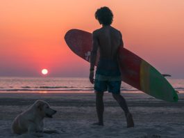 Man with surfboard stands on a Thai beach at sunset.