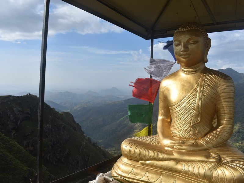 This image shows a golden Buddha with prayer flags strung behind. Behind the statue, there are tree-covered Sri Lankan mountains.